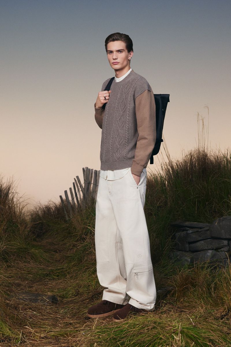 Male fashion model poses in a rural setting wearing a knit sweater, cream pants, and a backpack, capturing natural outdoor style.