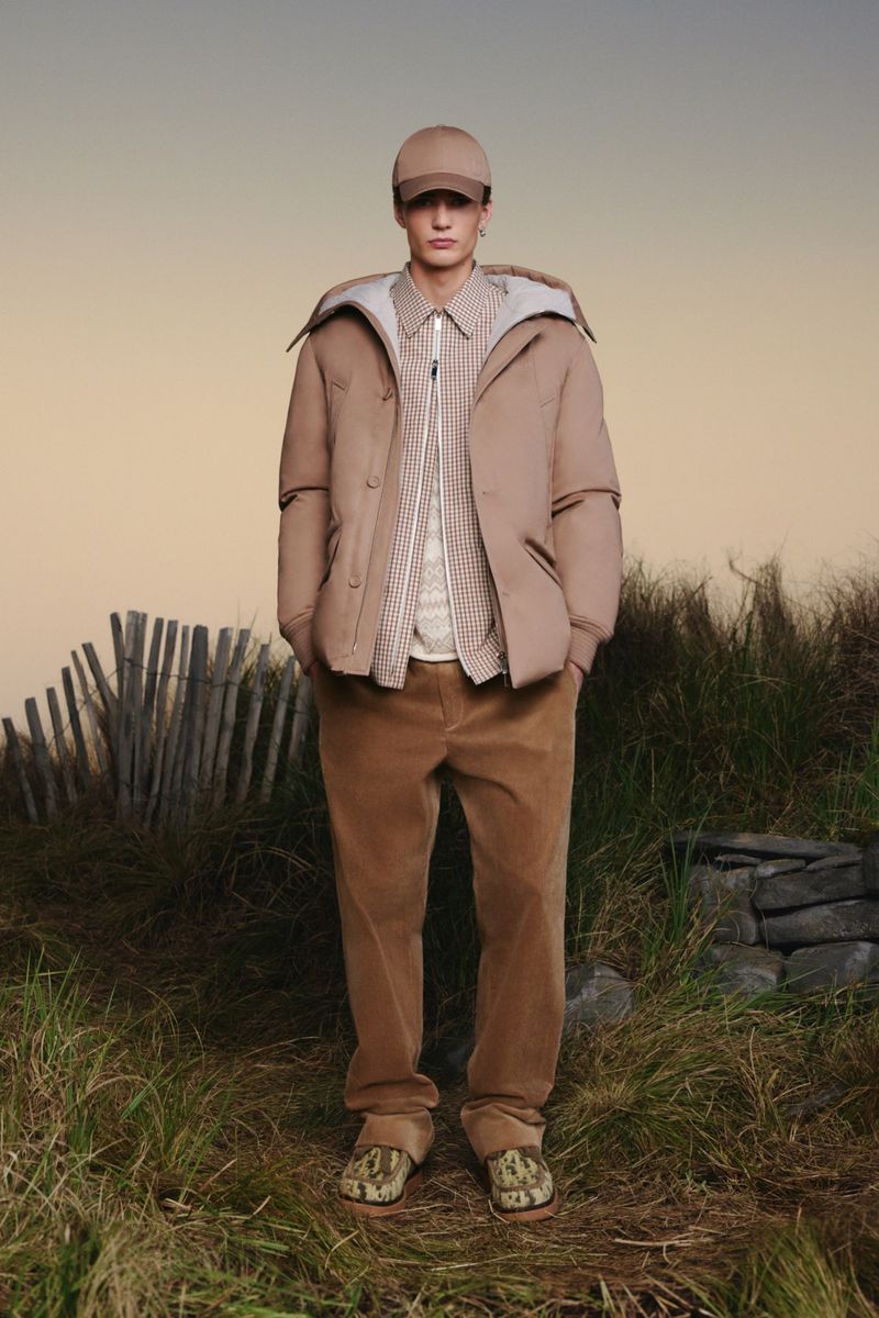 A full-length shot of a male fashion model in a cap, coat, patterned shirt, and pants standing in a grassy, outdoor environment.