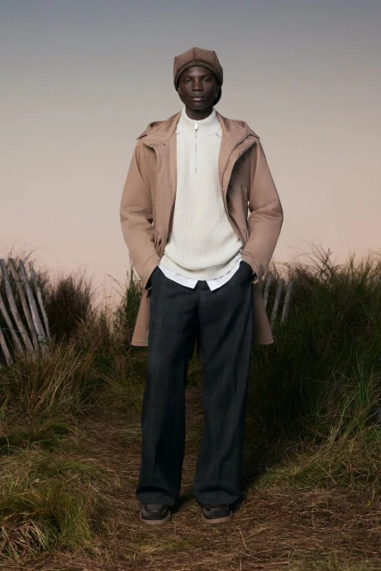 A Black male model stands outdoors on a grassy path, wearing a tan hooded raincoat, cream sweater, dark pants, brown cap, and brown shoes.