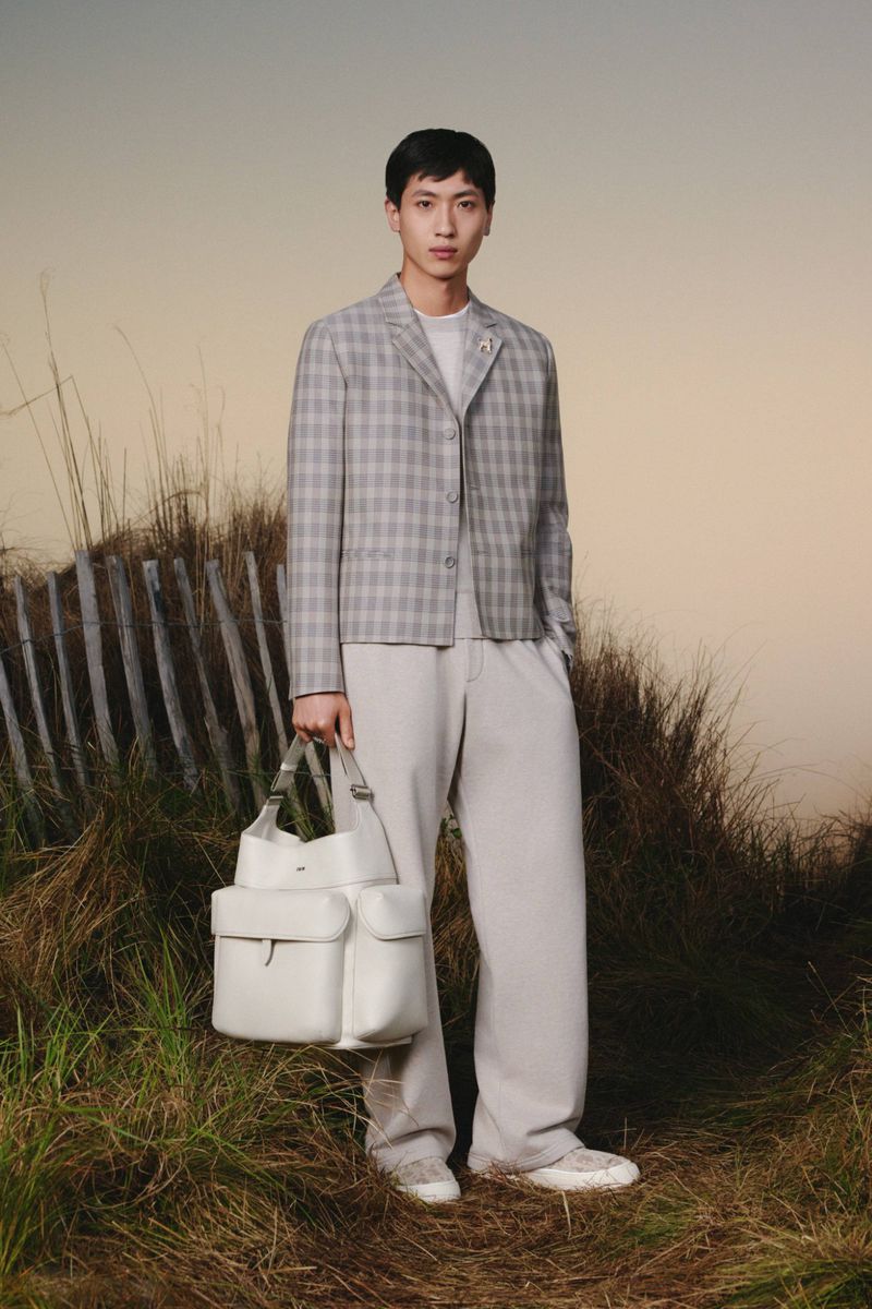 Male fashion model in plaid jacket and white pants holds a white leather bag, posing outdoors against a neutral sky background.
