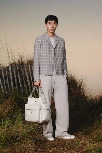 Male fashion model in plaid jacket and white pants holds a white leather bag, posing outdoors against a neutral sky background.