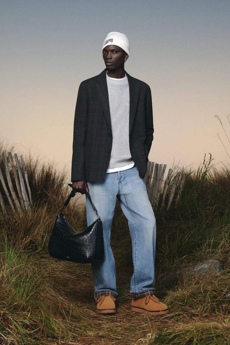 Male fashion model poses outdoors wearing a blazer, sweater, jeans, beanie, and carrying a leather bag in a fashion editorial style.