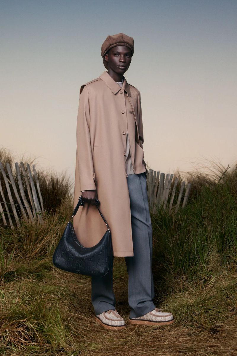 An African male model poses in a tan trench coat, beret, blue trousers, and patterned shoes while holding a black leather hobo bag.