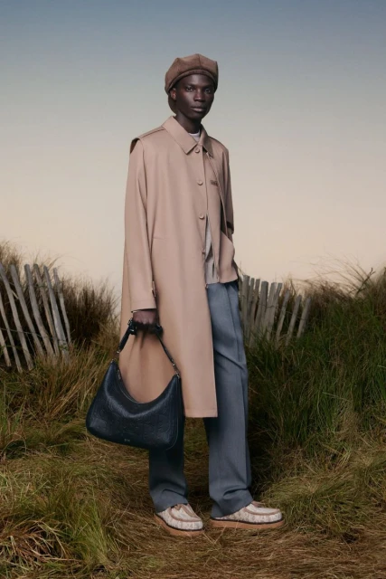 An African male model poses in a tan trench coat, beret, blue trousers, and patterned shoes while holding a black leather hobo bag.