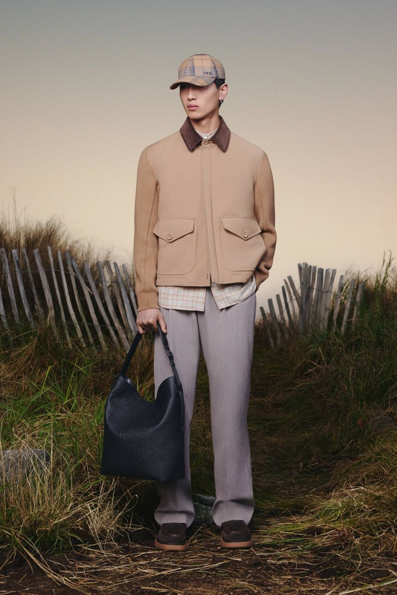 Asian male in neutral jacket and slacks poses with leather bag in outdoor setting with dunes and fence, overcast day, full shot.