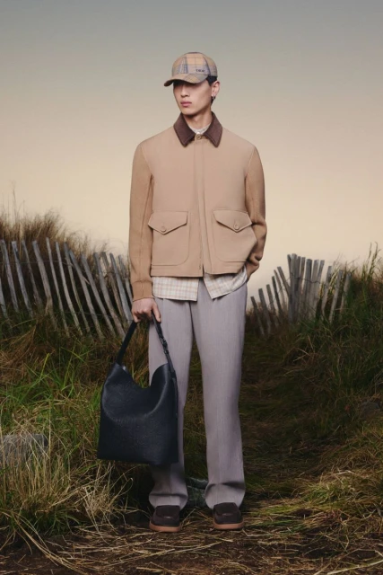 Asian male in neutral jacket and slacks poses with leather bag in outdoor setting with dunes and fence, overcast day, full shot.