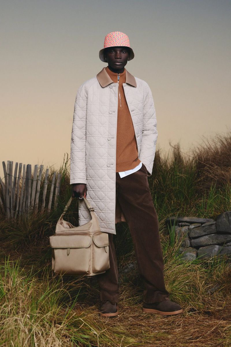 Dark-skinned man in bucket hat, quilted coat, tan sweater, brown pants, shoulder bag poses outdoors in natural lighting.