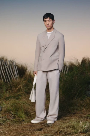 Man in a stylish double-breasted blazer and wide-leg pants, holding a handbag, poses outdoors against a soft, blurred natural background.