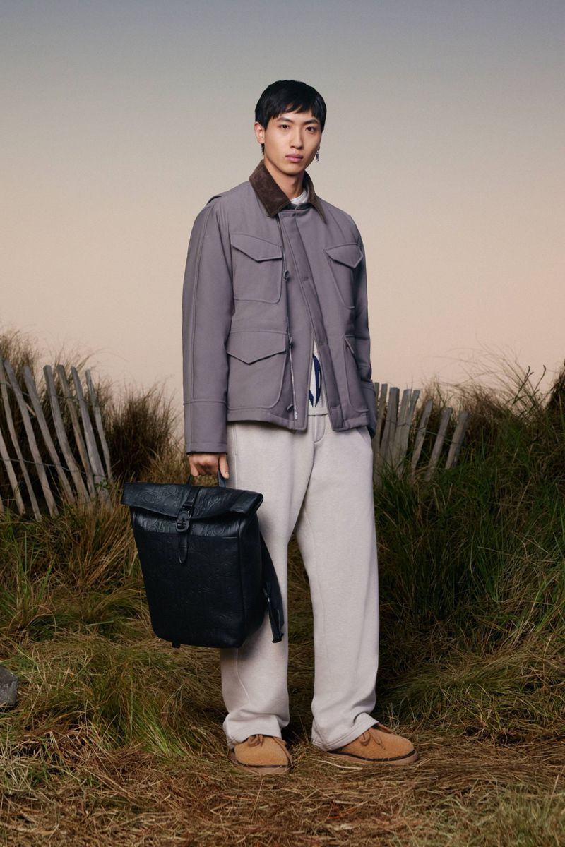Asian male model poses in nature holding a dark backpack wearing a gray jacket and tan pants in a medium, eye-level shot.