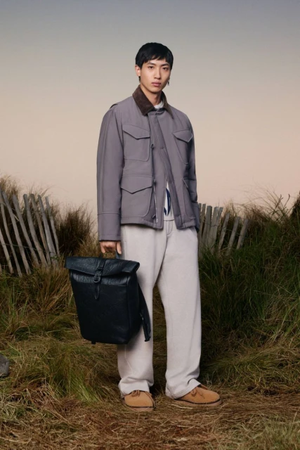 Asian male model poses in nature holding a dark backpack wearing a gray jacket and tan pants in a medium, eye-level shot.