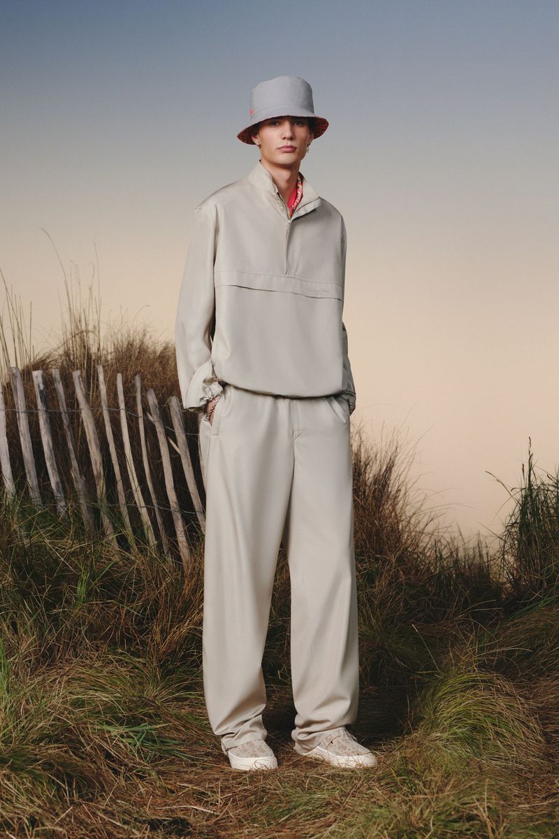 A young man models beige streetwear: bucket hat, anorak, pants, and sneakers, standing outdoors in a grassy, natural environment.