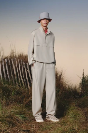A young man models beige streetwear: bucket hat, anorak, pants, and sneakers, standing outdoors in a grassy, natural environment.