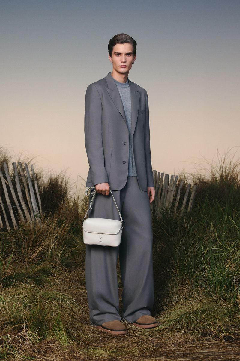 Male model in a grey suit and sweater stands outdoors holding a cream-colored bag, on a grassy path with a fence in the background.