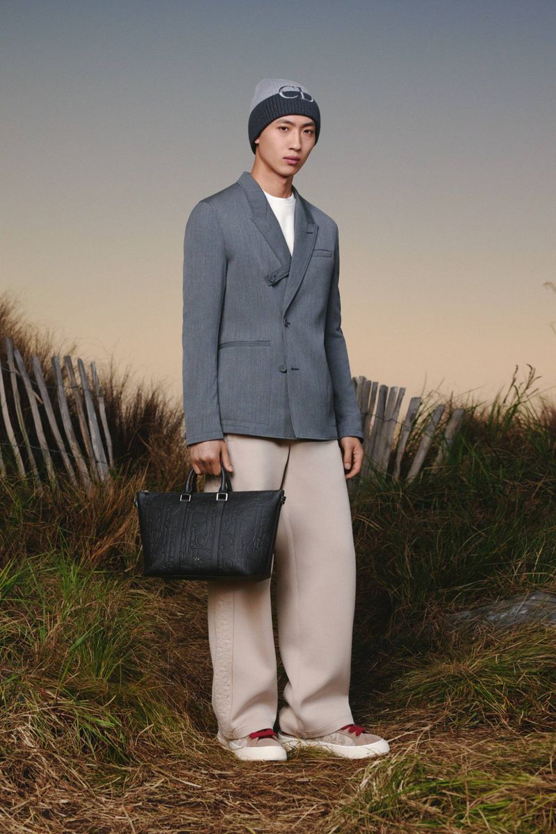 An Asian male model poses outdoors in a gray blazer, beige pants, and a beanie, holding a black tote bag, against a blurred background.