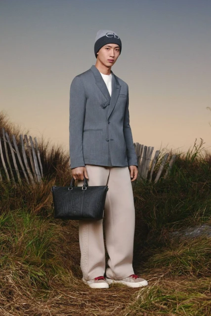 An Asian male model poses outdoors in a gray blazer, beige pants, and a beanie, holding a black tote bag, against a blurred background.