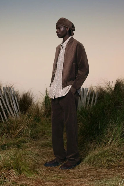 A black male model poses in a brown corduroy outfit—jacket, pants, and beret—on a grassy field with a cloudy sky backdrop, fashion shot.