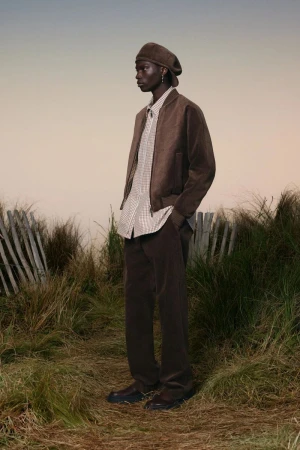 A black male model poses in a brown corduroy outfit—jacket, pants, and beret—on a grassy field with a cloudy sky backdrop, fashion shot.