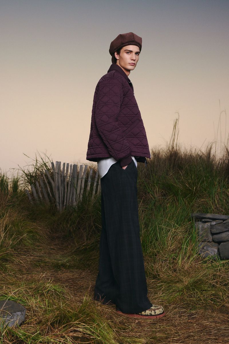 A young man with a beret and a quilted jacket poses outdoors in a field. He is wearing dark pants and colorful shoes.