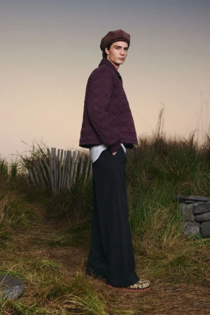 A young man with a beret and a quilted jacket poses outdoors in a field. He is wearing dark pants and colorful shoes.