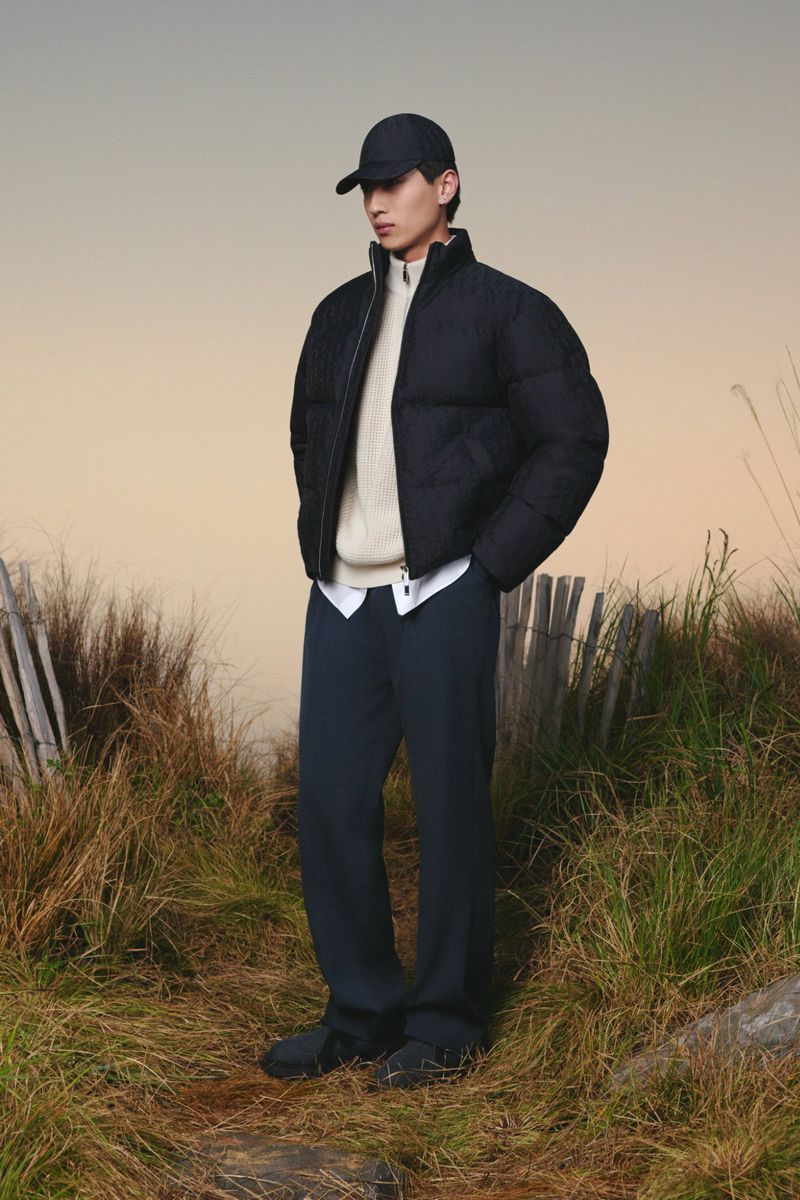 A fashionable adult male poses in a black puffer jacket and navy trousers outdoors surrounded by grassy vegetation in a lifestyle shot.