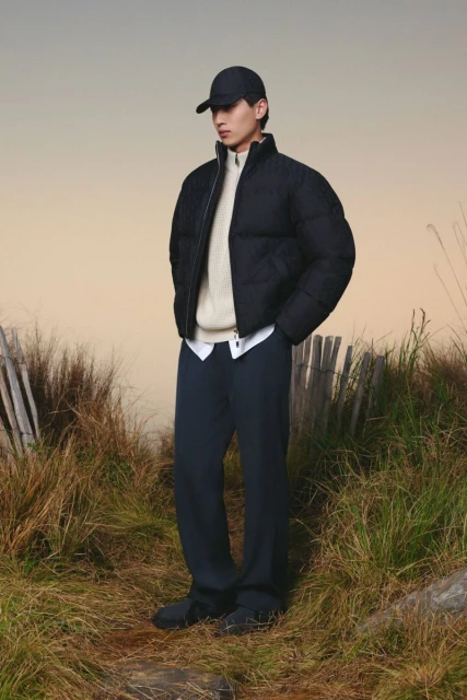 A fashionable adult male poses in a black puffer jacket and navy trousers outdoors surrounded by grassy vegetation in a lifestyle shot.