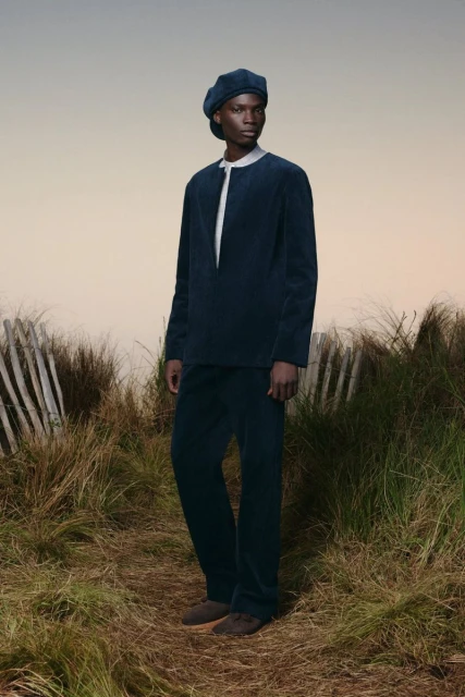 Adult male fashion model posing outdoors in blue corduroy shirt, pants, and beret on a grassy path with a beige sky background.