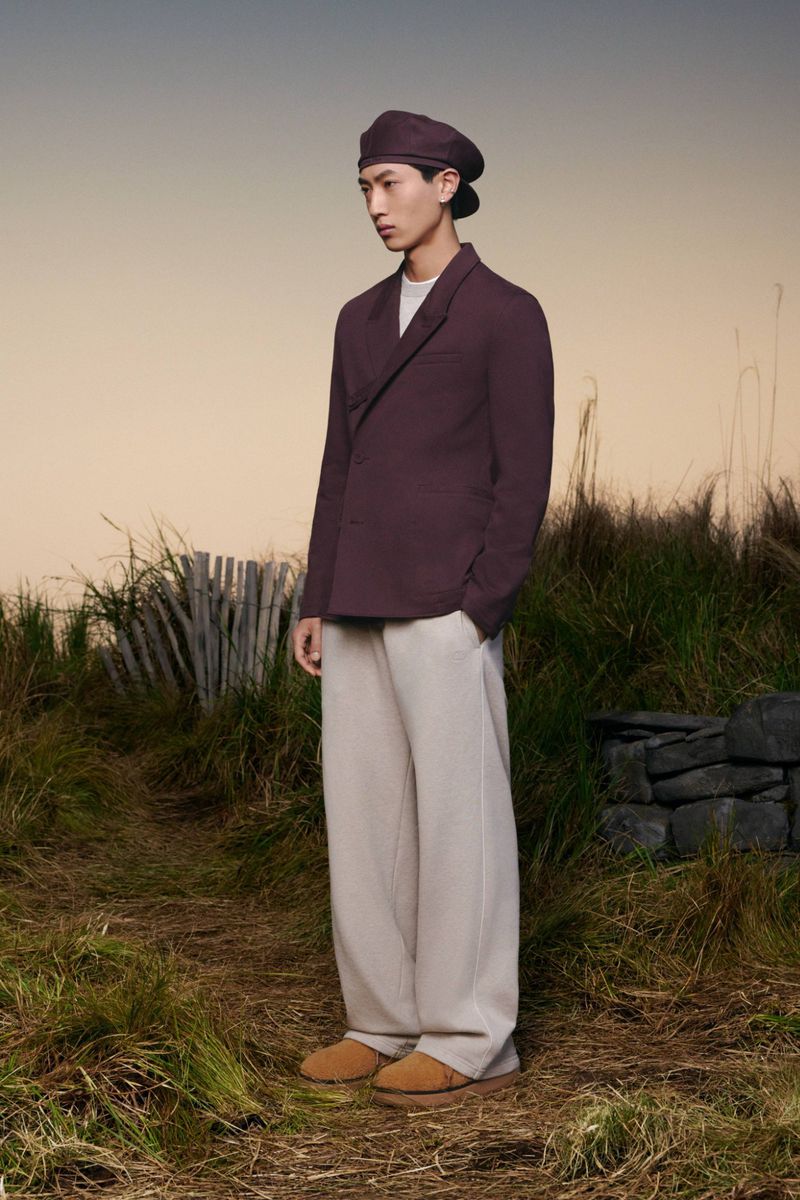 Full-length shot of a person posing in a blazer, wide-legged pants, and a beret on a grassy field with a neutral sky backdrop.
