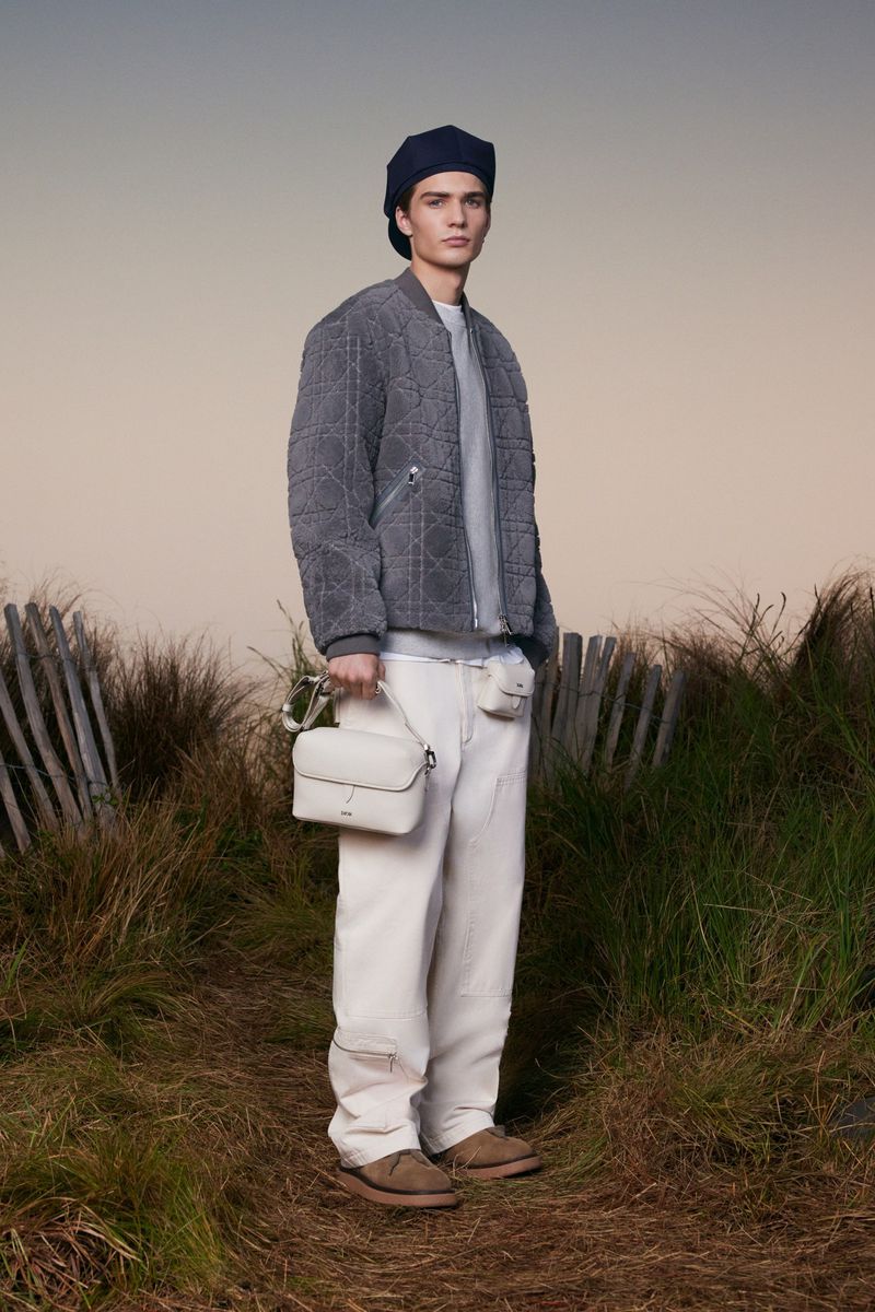 Model in a grey quilted bomber jacket, white trousers, and dark beret holding a cream leather bag outdoors in grassy dunes.