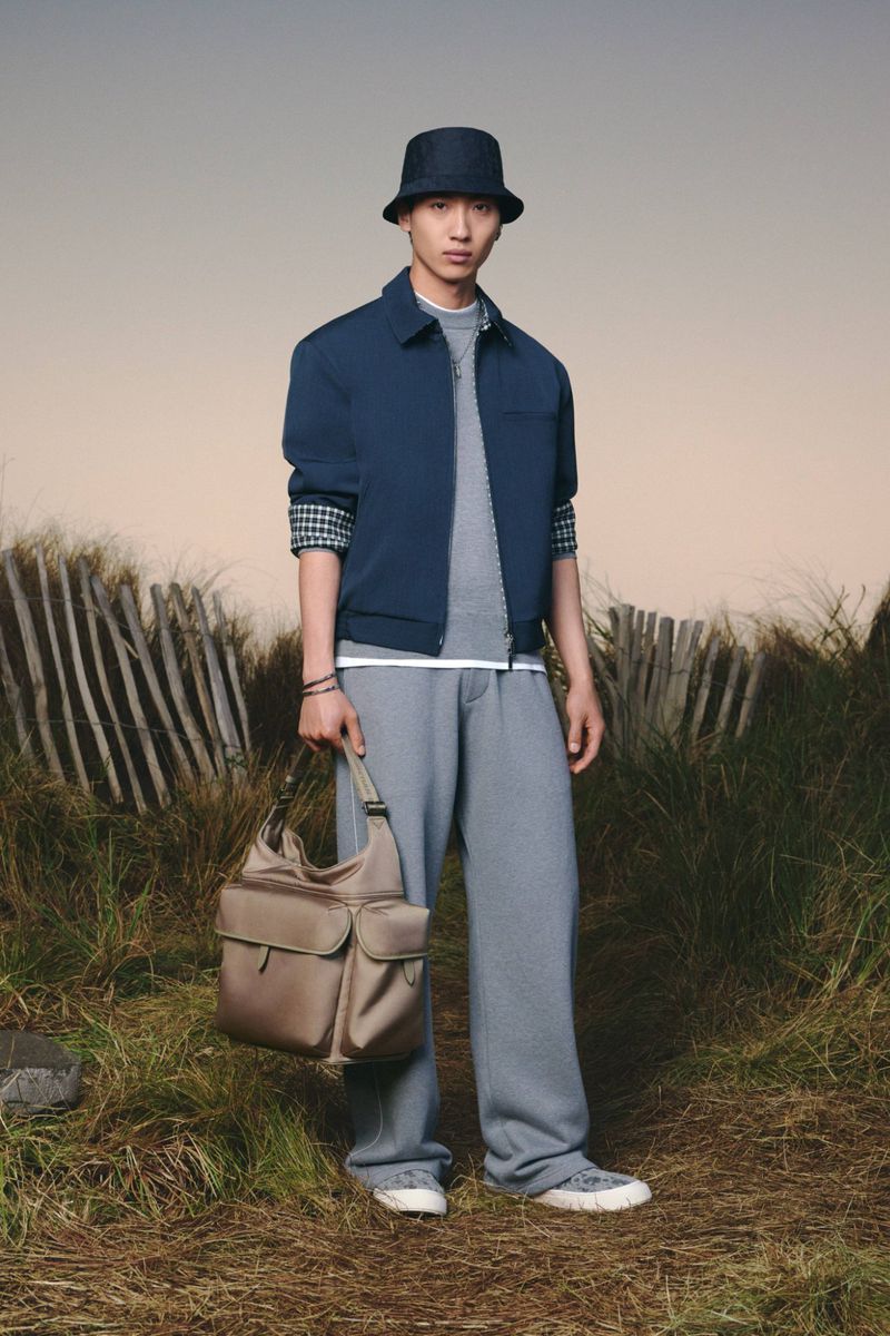 Asian male fashion model poses outdoors wearing a blue jacket, gray pants, and bucket hat, holding a satchel bag in a grassy field.