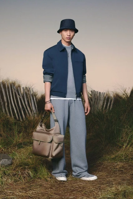 Asian male fashion model poses outdoors wearing a blue jacket, gray pants, and bucket hat, holding a satchel bag in a grassy field.