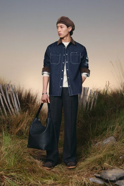 Man posing outdoors with a denim shirt, black trousers, and a large leather handbag, standing in a grassy field with a fence.