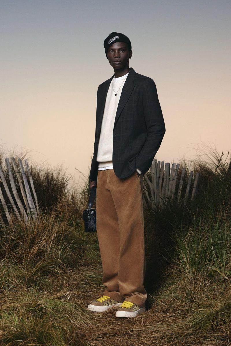 Dark-skinned male model in blazer and corduroy pants stands outdoors in tall grass against a dusky sky wearing a dark cap.