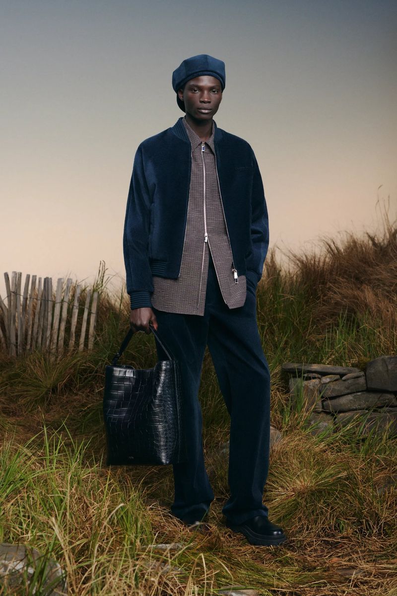 Model Dara Gueye standing in a coastal grassy field wearing a navy jacket, matching trousers, and beret with a black leather tote bag.