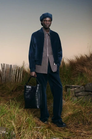Model Dara Gueye standing in a coastal grassy field wearing a navy jacket, matching trousers, and beret with a black leather tote bag.