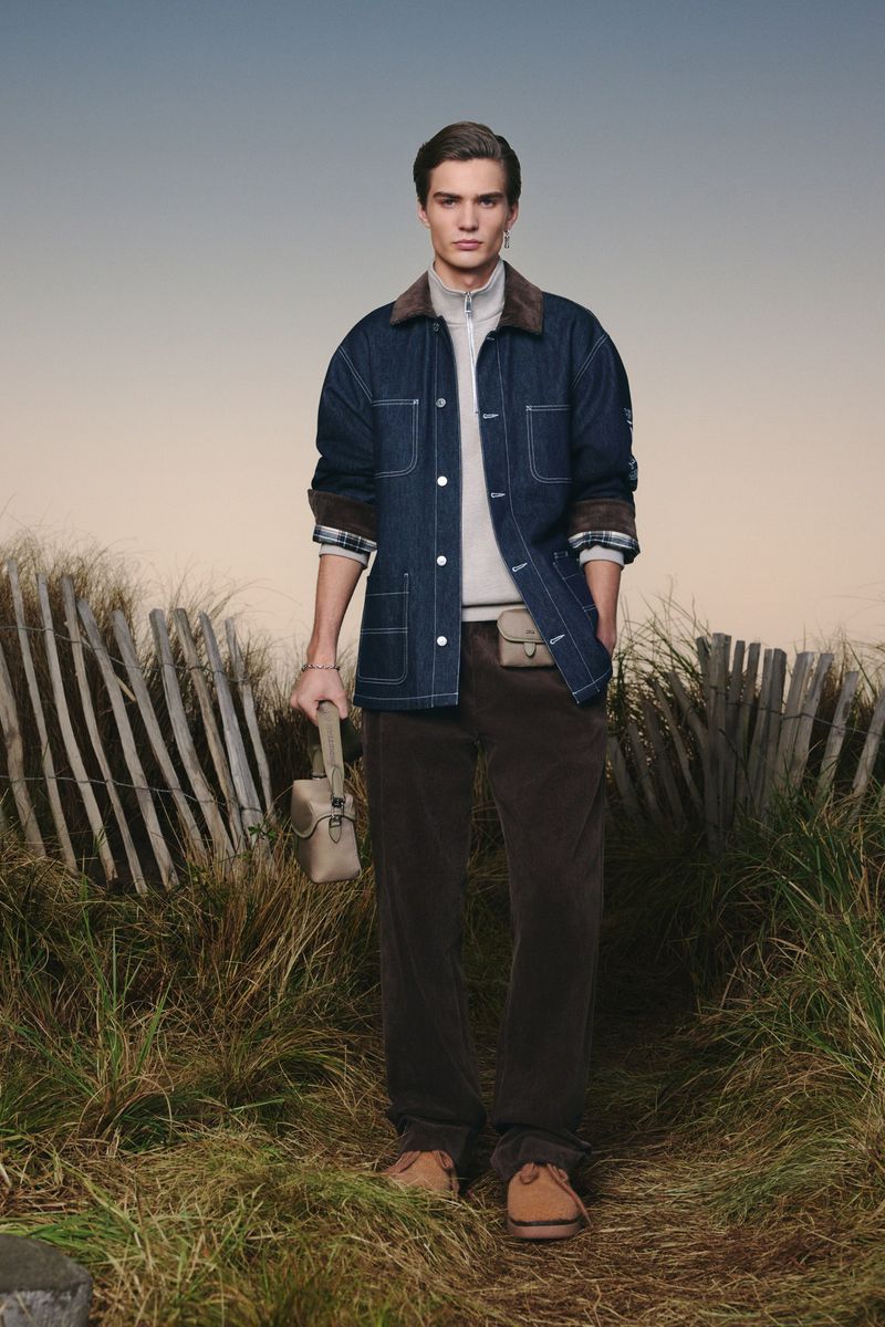 Man stands in outdoor fashion shot. He wears a blue denim jacket with brown corduroy collar, brown pants, and carries a small bag.
