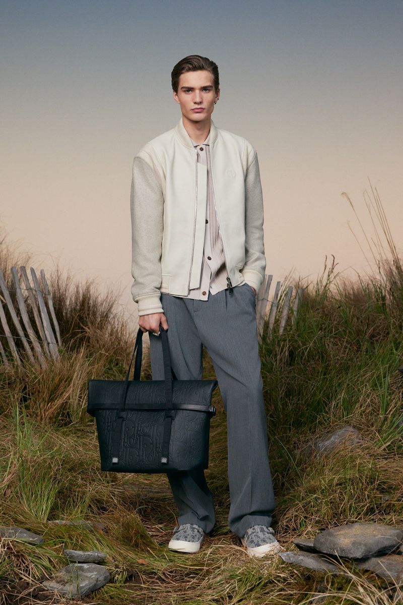 Man in light jacket and grey pants stands in field, holding black leather tote bag against soft, diffused lighting and natural backdrop.