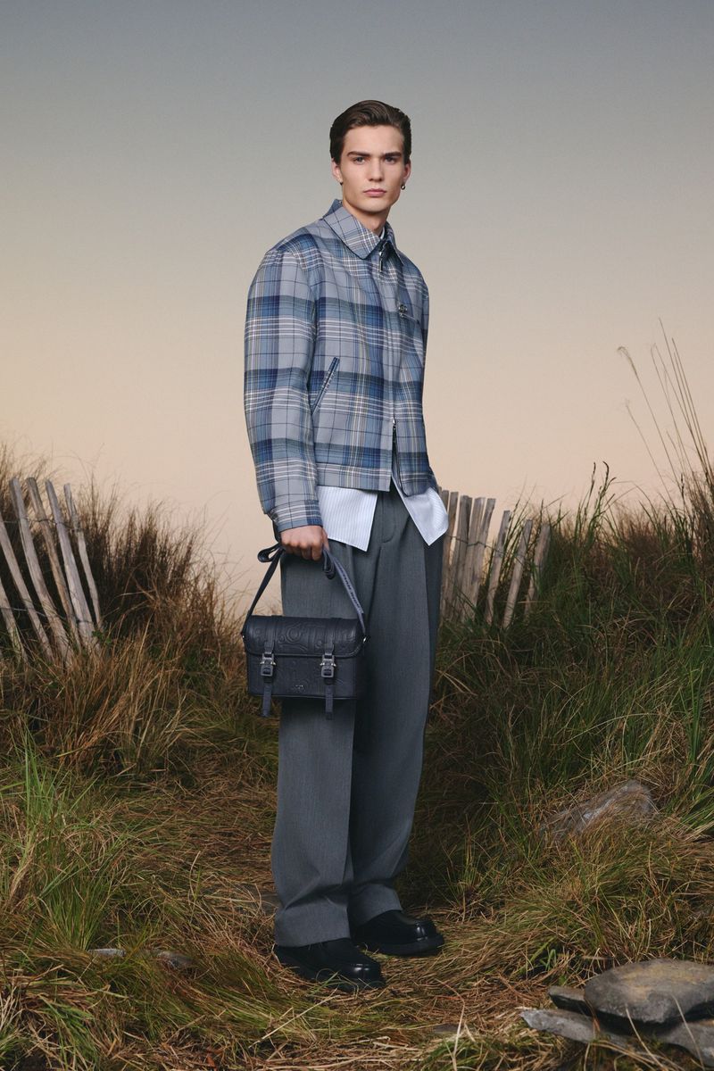 Man in gray plaid jacket and wide leg pants holding black leather satchel bag in an outdoor field fashion portrait.