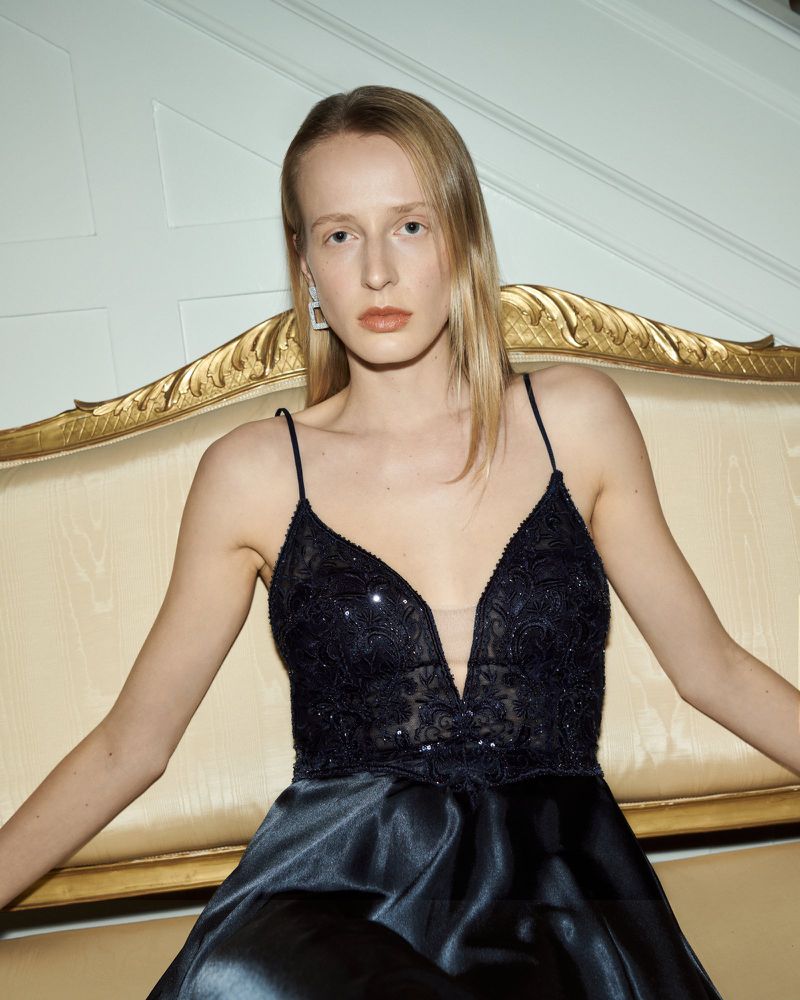Elegant woman in a formal black gown posing indoors. The dress features a low V-neck and a satin skirt, with a cream colored background.