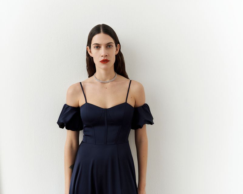 Portrait of a woman wearing a navy blue dress with an off-the-shoulder neckline against a white wall in a photo studio setting.