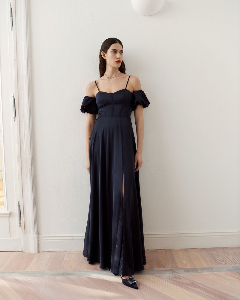 Elegant woman in navy gown with shoulder straps and slit standing in bright neutral interior with wood floors by window.