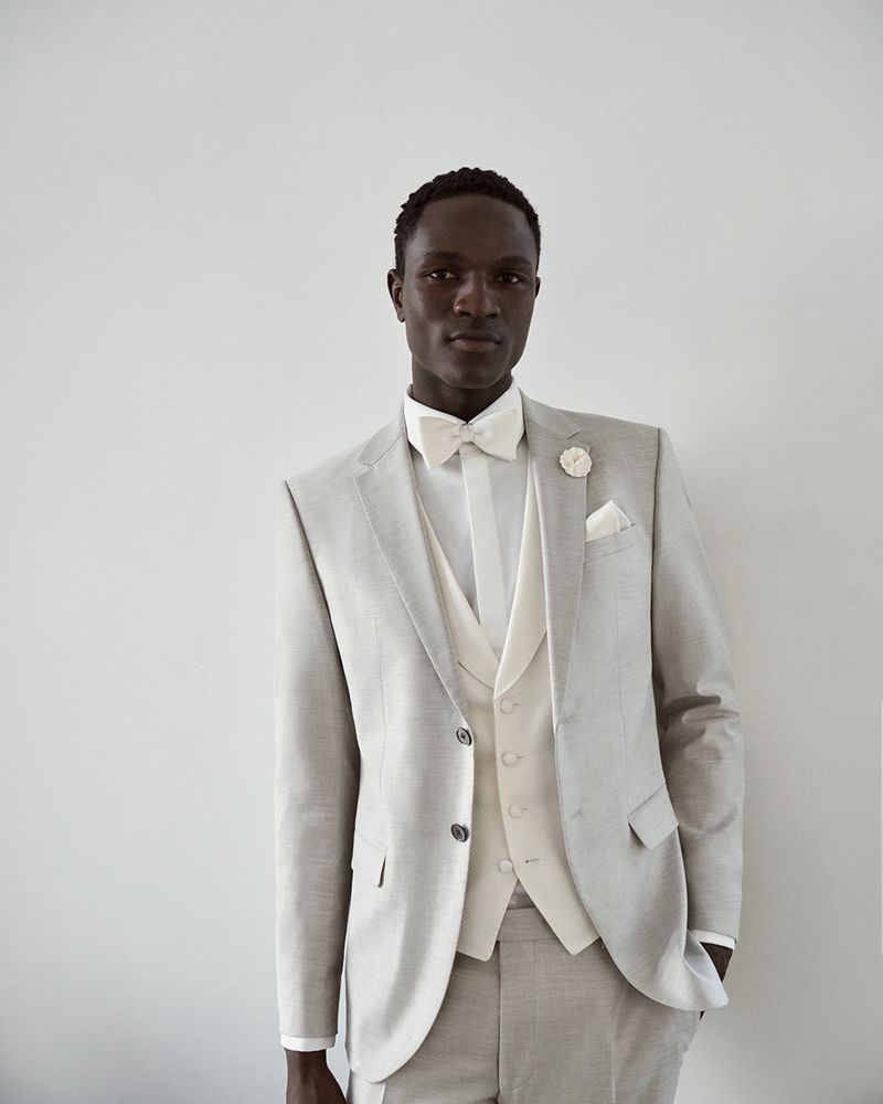 A single Black male model in a light grey three-piece suit with a bow tie poses against a neutral background for a fashion editorial.