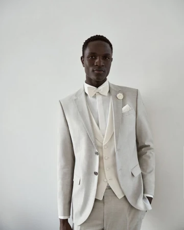 A single Black male model in a light grey three-piece suit with a bow tie poses against a neutral background for a fashion editorial.