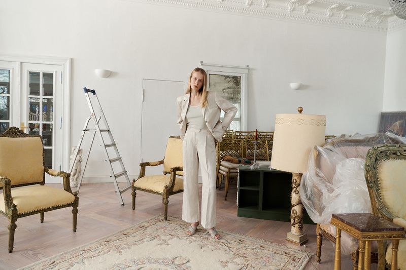 Fashion shot of a blonde woman in a light suit standing indoors with furniture and a ladder, in a bright, sparsely decorated room.