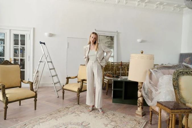 Model Anine Van Velzen in a cream tailored pantsuit standing in a room with vintage yellow armchairs and a stepladder.