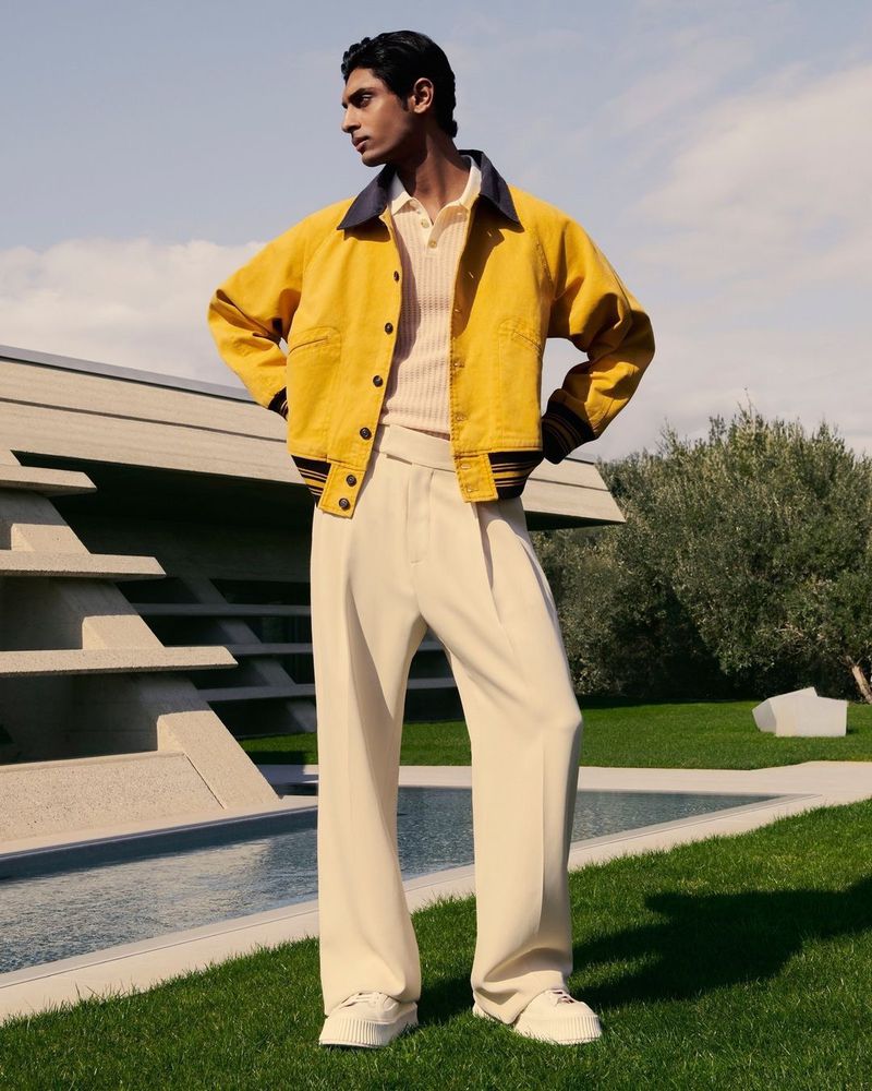 A fashionable adult male poses in a bright yellow jacket and wide white trousers, set against a modern architectural backdrop and green lawn.