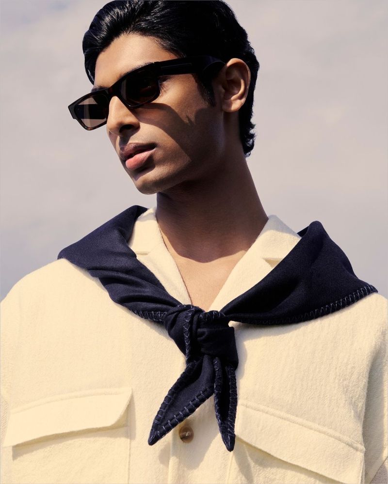 Portrait of a young man wearing sunglasses and a knotted navy scarf, posing in natural light against a bright sky background.