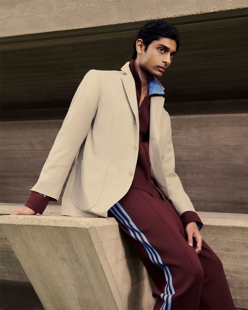 Fashionable adult male in a beige blazer and maroon pants with light blue stripes, posing against a raw concrete structure backdrop.