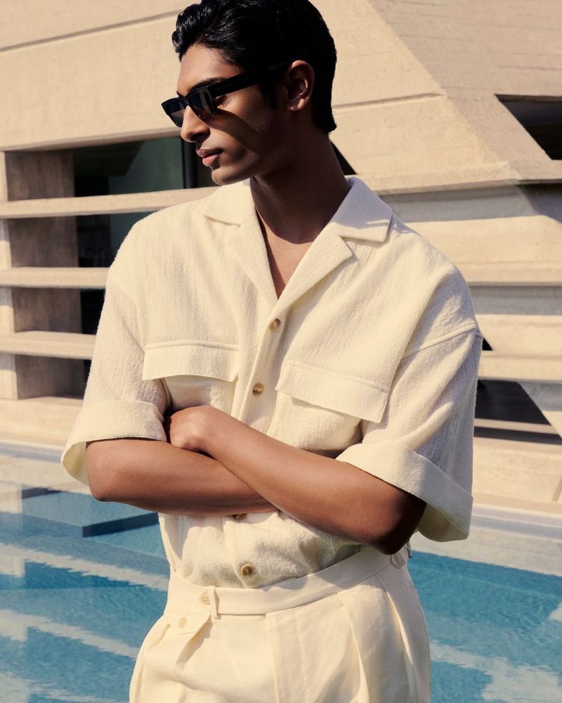 Pratik Shetty wearing a cream short-sleeved shirt and black sunglasses by an outdoor pool for Mytheresa Men.
