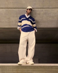 Model Pratik Shetty standing in a blue and cream striped sweater, wide-leg trousers, and cap against a concrete wall.
