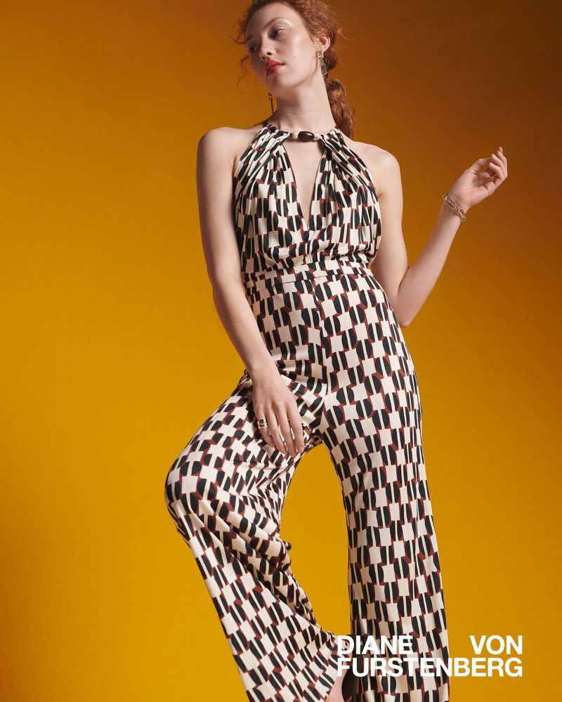 A stylish woman poses in a patterned Diane von Furstenberg jumpsuit against a vibrant orange background, showcasing contemporary fashion.